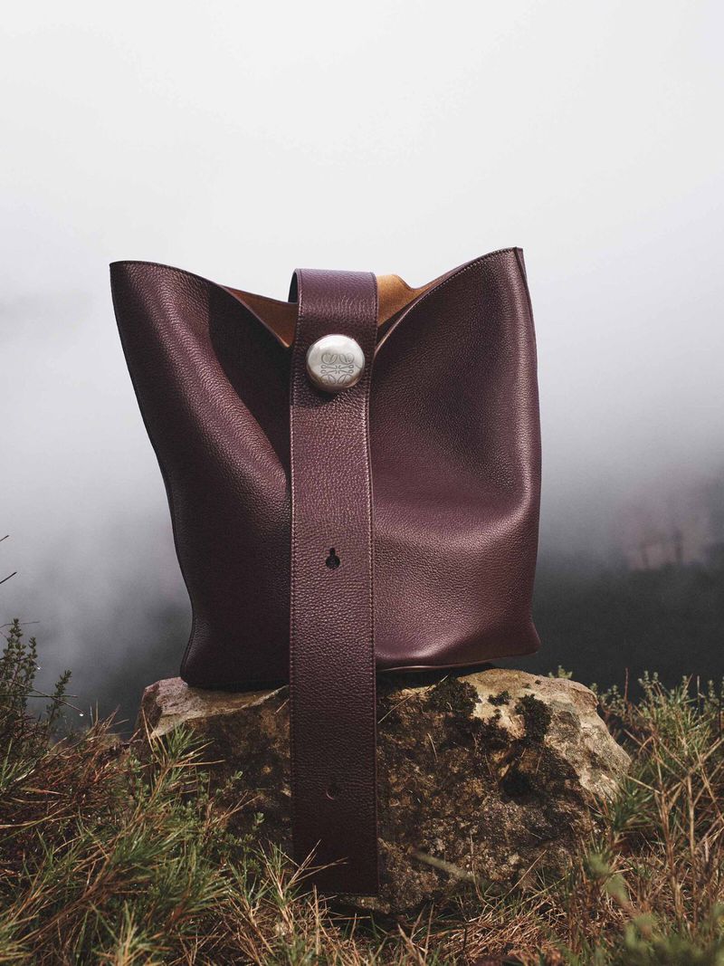 Burgundy leather bucket bag with a silver button sits on a mossy rock, photographed outdoors with a misty forest background.