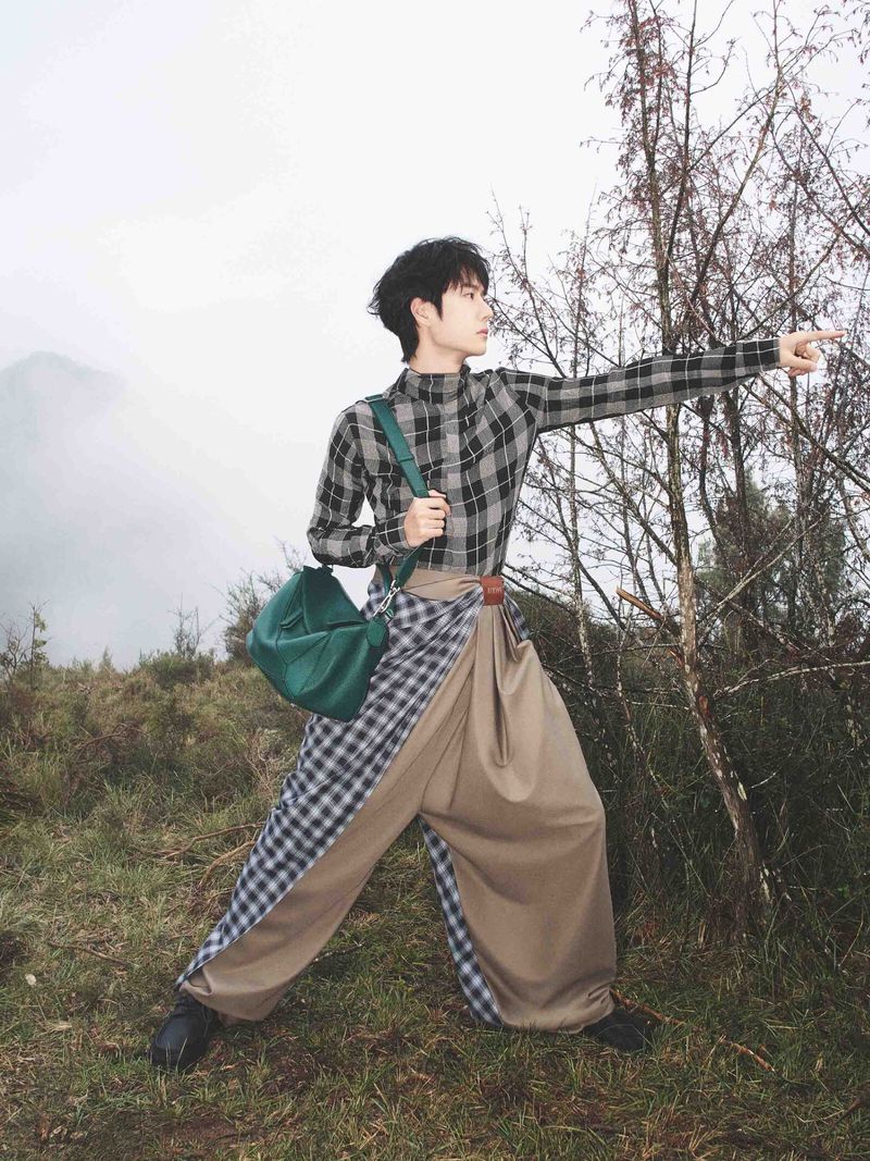 Man wearing a plaid shirt and draped beige trousers pointing forward outdoors holding a green handbag