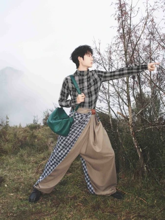 Man wearing a plaid shirt and draped beige trousers pointing forward outdoors holding a green handbag