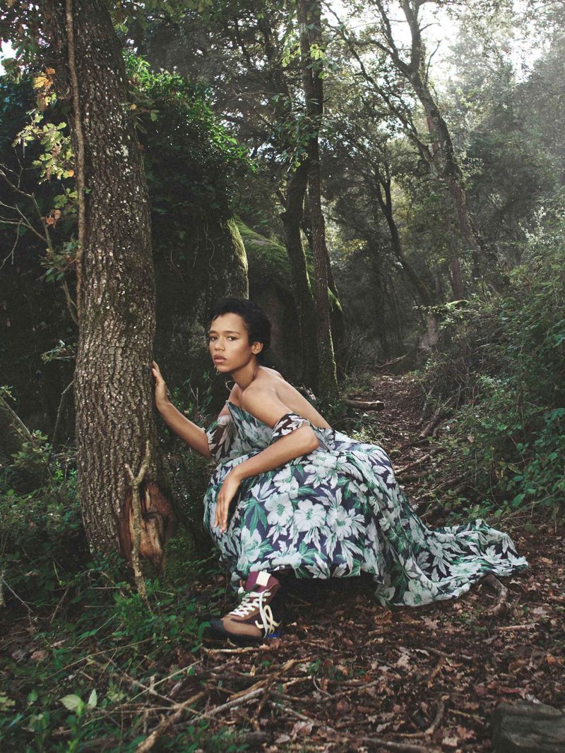 Young woman in a flowing floral dress poses in a forest with tall trees and a dirt path, fashion and nature converge in this scene.
