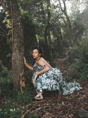 Model in a floral print off-the-shoulder dress crouching beside a tree trunk in a lush green forest.
