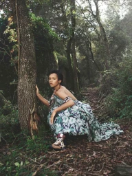 Model in a floral print off-the-shoulder dress crouching beside a tree trunk in a lush green forest.