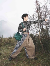 Man wearing a plaid shirt and draped beige trousers pointing forward outdoors holding a green handbag