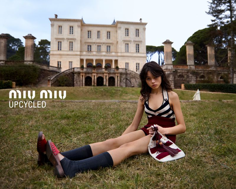Brunette woman in striped top and burgundy skirt sits on lawn with bag and building in background for a fashion editorial.