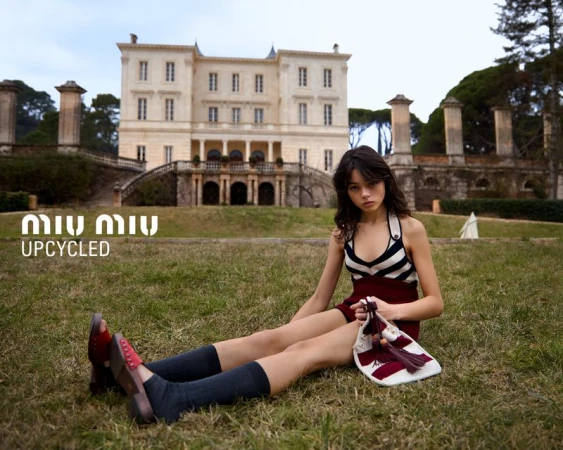Brunette woman in striped top and burgundy skirt sits on lawn with bag and building in background for a fashion editorial.