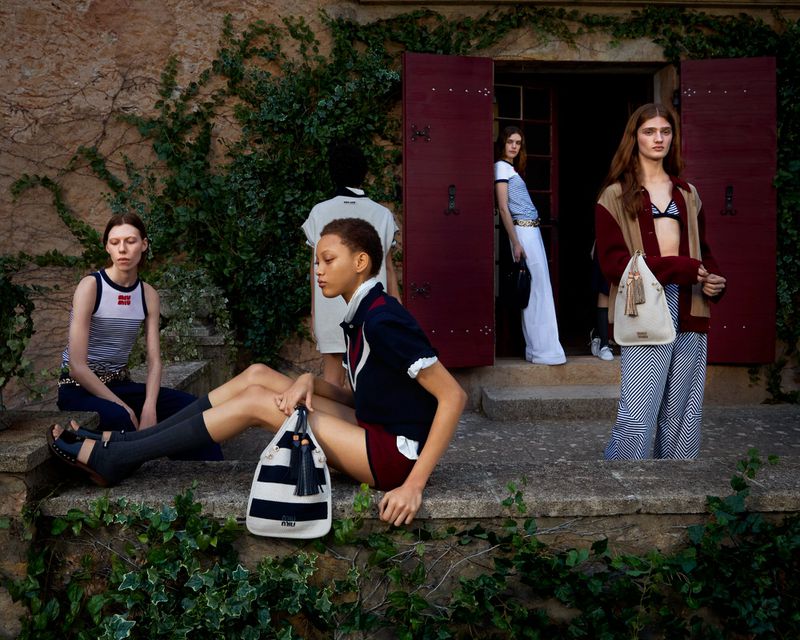 Fashion shoot of five women in matching preppy-chic outfits standing on the steps of a building with red doors and green vines.