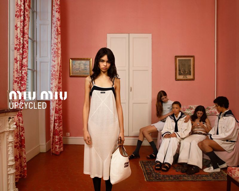 Fashion editorial photo: a slender woman poses in a light dress, with other models lounging on a sofa in a pink-toned room.