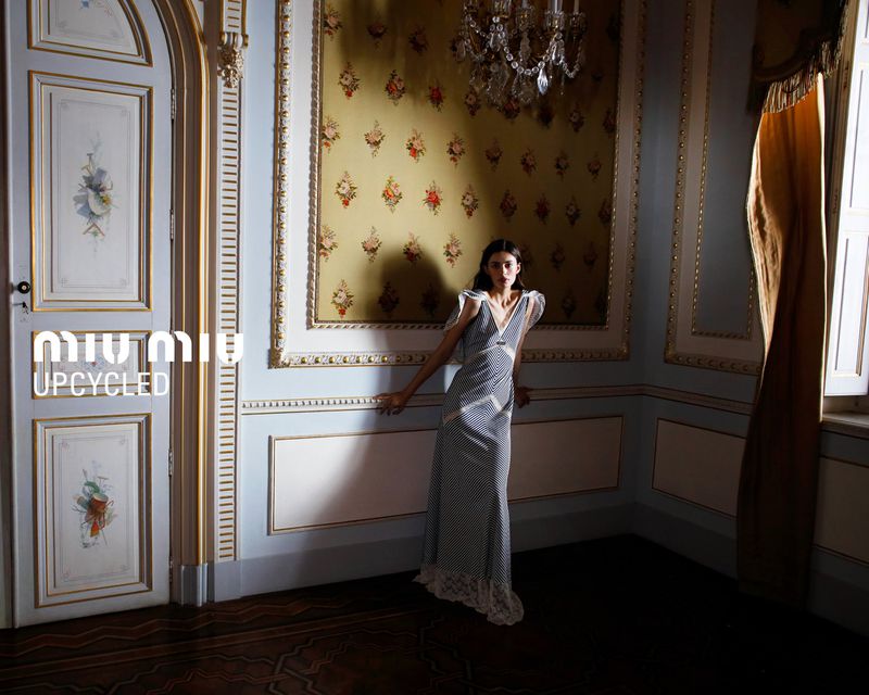 Diana Silvers posing in a long printed slip dress with lace trim against ornate floral wallpaper in a classical interior room.