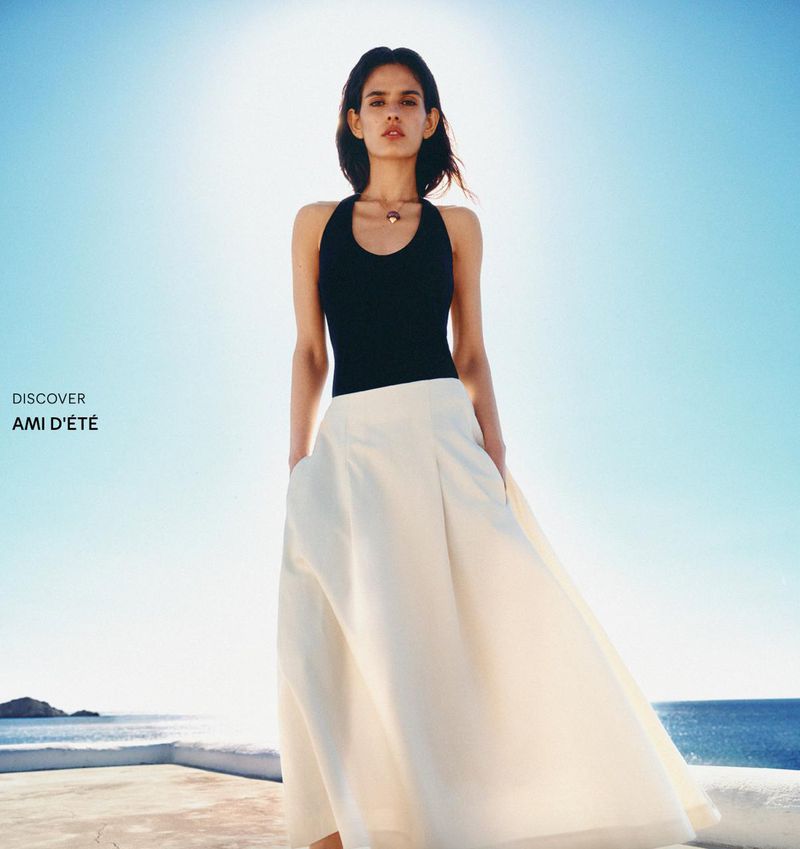 Avishag Amir wears a black halter top and cream maxi skirt against a bright blue coastal sky for Ami D'Été.