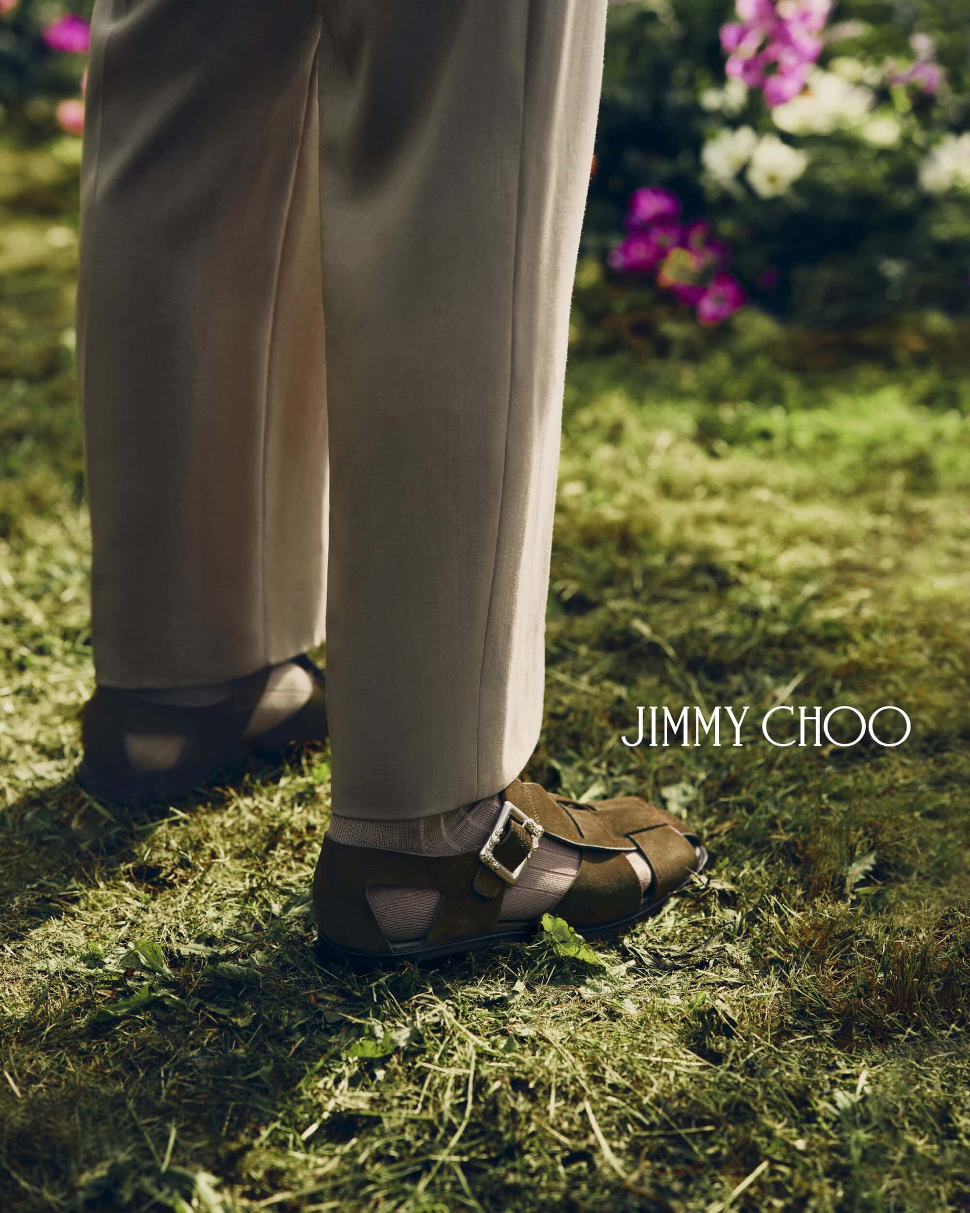 Model wearing beige tailored trousers and olive suede buckle sandals standing on grass for Jimmy Choo.
