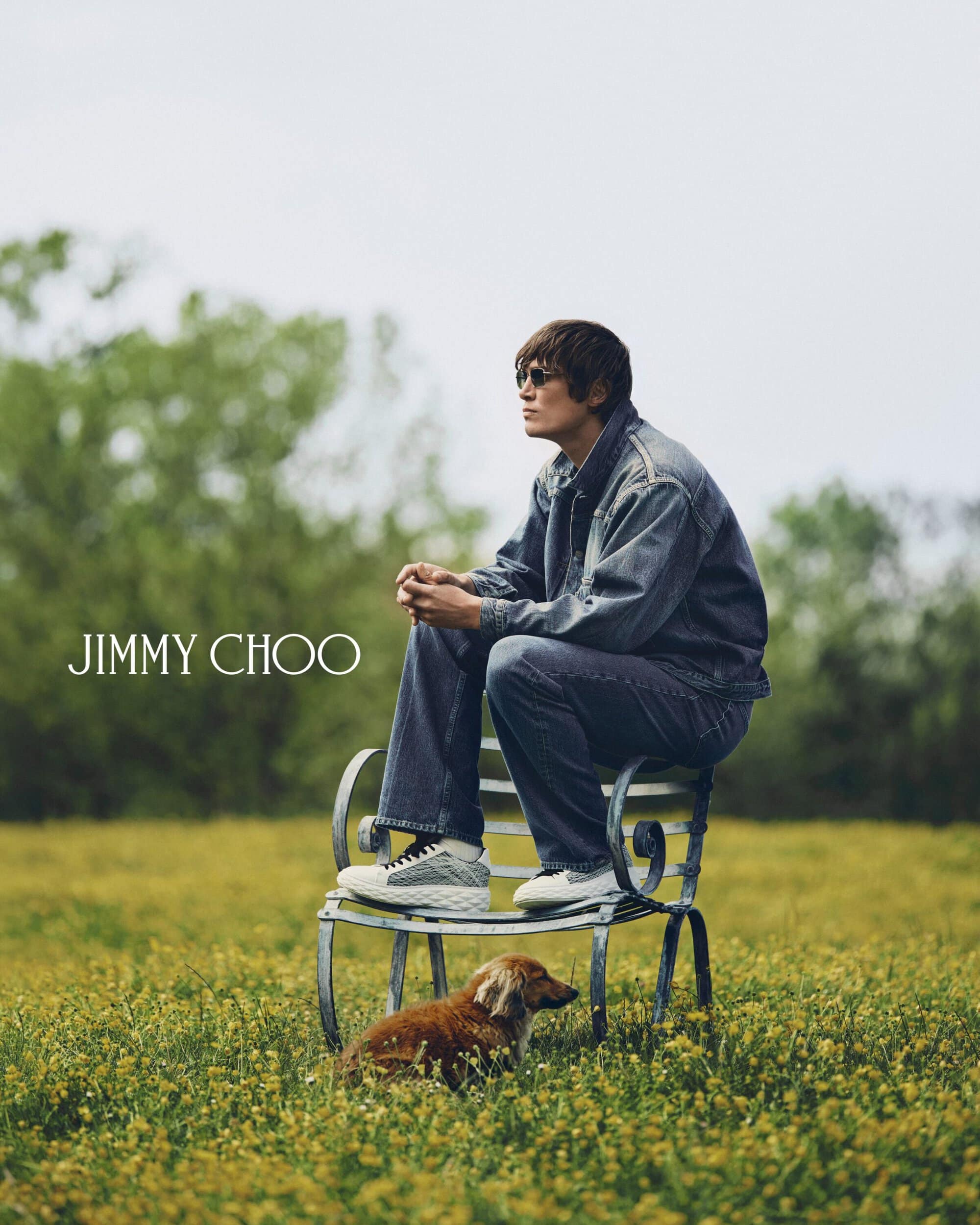 Male model wearing denim jacket and jeans seated on a wrought-iron bench with a small dog on a grassy meadow under an overcast sky