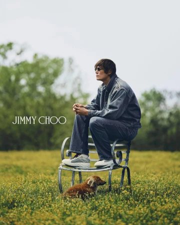 Male model wearing denim jacket and jeans seated on a wrought-iron bench with a small dog on a grassy meadow under an overcast sky