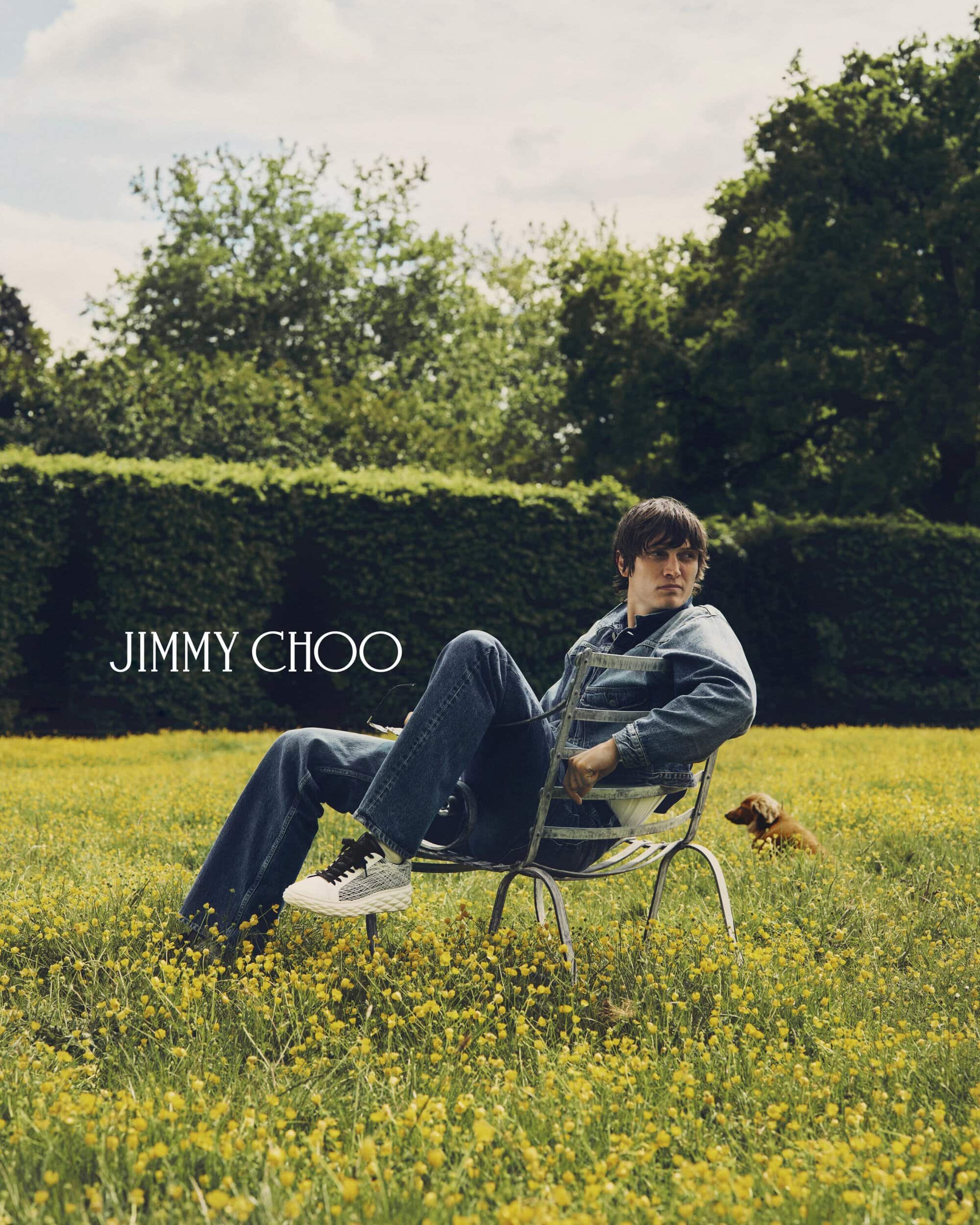 Man seated on a metal chair in a yellow meadow wearing denim jacket and jeans, with a small dog in the background.