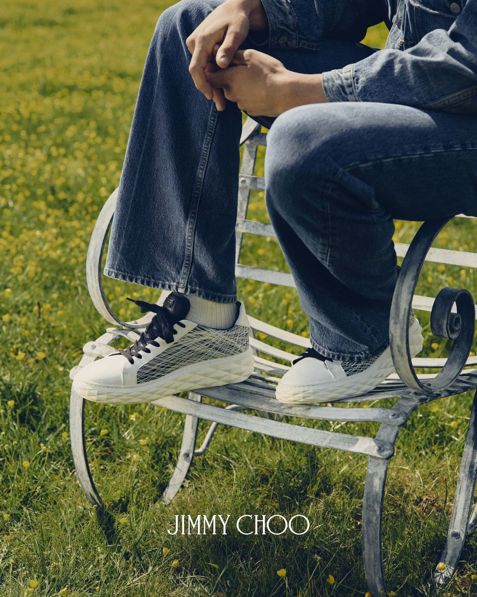 Model seated on a white metal bench wearing blue jeans and textured Jimmy Choo sneakers in a grassy field.