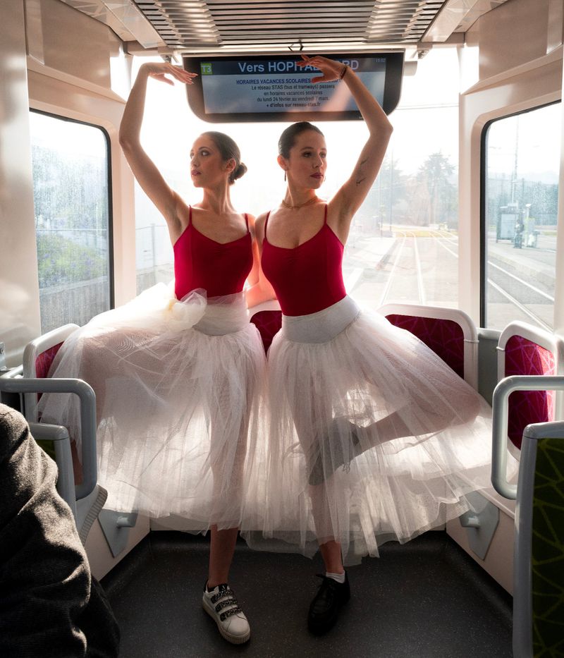 Two ballerinas in red leotards and white tutus strike a pose inside a train car near windows, illuminated by natural light.