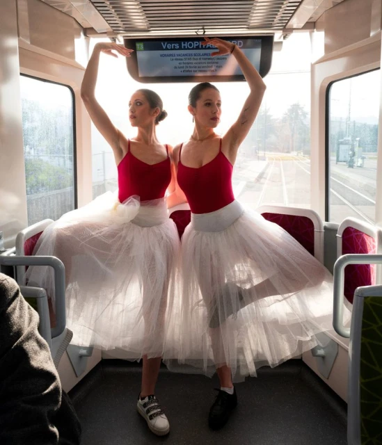 Two ballerinas in red leotards and white tutus strike a pose inside a train car near windows, illuminated by natural light.
