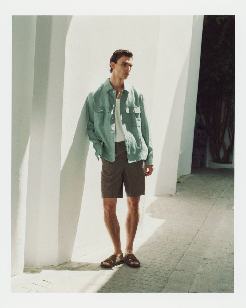 Male model leaning against a white architectural wall wearing a light green overshirt, white tank top, and dark patterned shorts.
