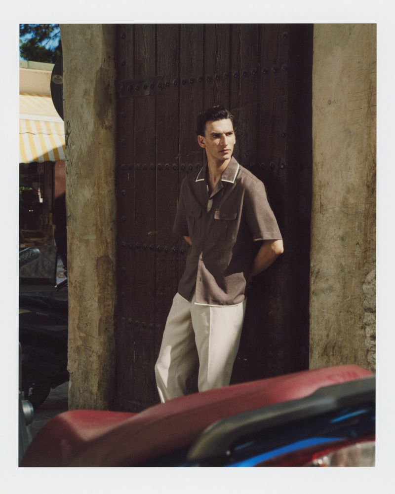 Model Thibaud Charon leaning against a rustic wooden door wearing a brown short-sleeved collared shirt and cream trousers.