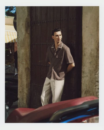 Model Thibaud Charon leaning against a rustic wooden door wearing a brown short-sleeved collared shirt and cream trousers.