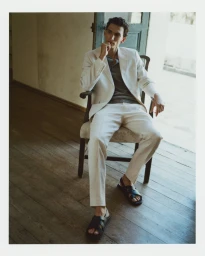 Model Thibaud Charon seated in a vintage wooden armchair wearing a cream linen suit and black leather sandals.