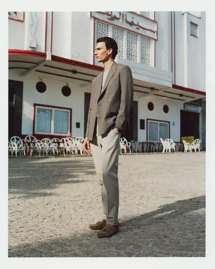 Model Thibaud Charon wearing a taupe double-breasted blazer and light trousers standing on a cobblestone plaza in front of a white building.