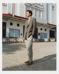 Model Thibaud Charon wearing a taupe double-breasted blazer and light trousers standing on a cobblestone plaza in front of a white building.