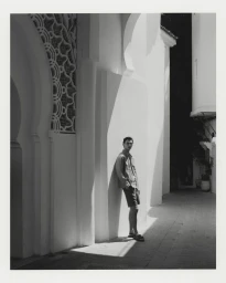 Model Thibaud Charon leaning against a sunlit white architectural wall in an alley wearing a light jacket, shorts, and sandals.