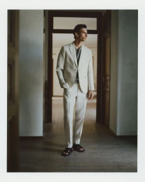 Model Thibaud Charon standing in an interior doorway wearing an off-white linen suit, polo shirt, and black sandals.