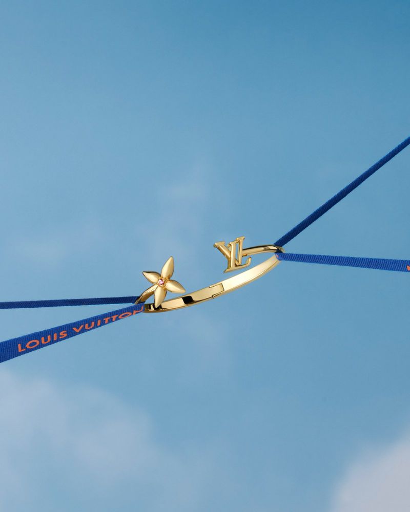 Gold-tone Louis Vuitton bracelet featuring a floral design and monogram, suspended by blue ribbons against a light blue background.