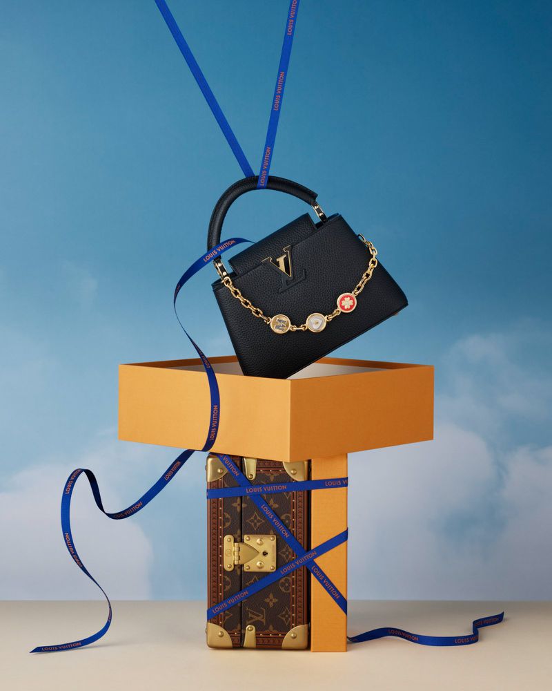 Luxury black leather Louis Vuitton bag with gold chain detail atop an orange box and vintage trunk, tied with a blue ribbon. 