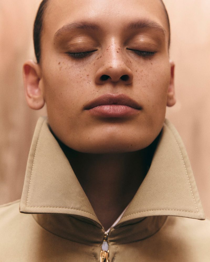 Close-up portrait of a freckled model with eyes closed wearing a beige high-collar zippered jacket against a warm backdrop.