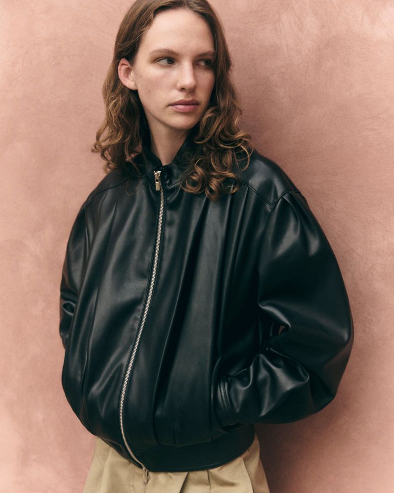 Model wearing an oversized black leather bomber jacket and beige pleated trousers against a textured pink background.