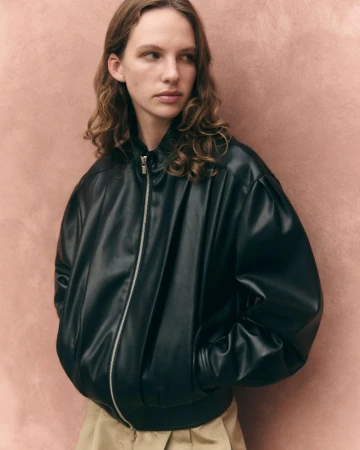Model wearing an oversized black leather bomber jacket and beige pleated trousers against a textured pink background.