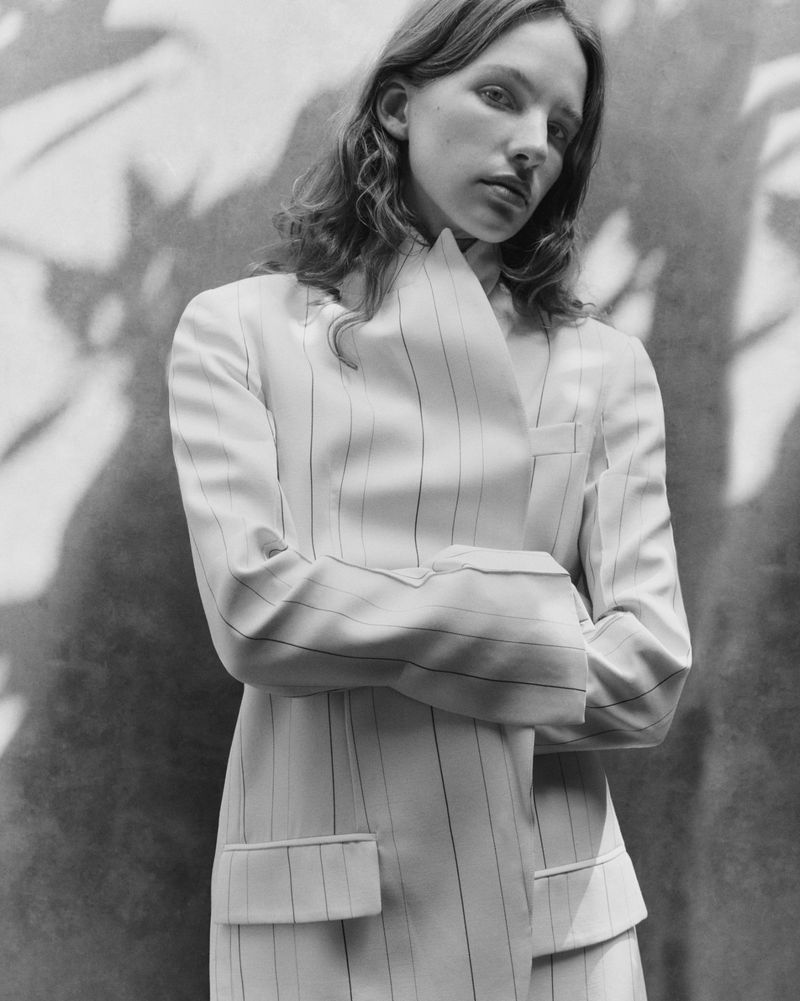 Model with folded arms wearing a structured white pinstripe blazer in a black and white portrait with leaf shadows.