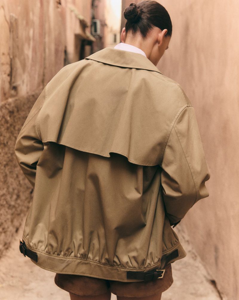 A model with hair in a bun seen from behind, wearing an oversized khaki storm-flap jacket while standing in a narrow alleyway.