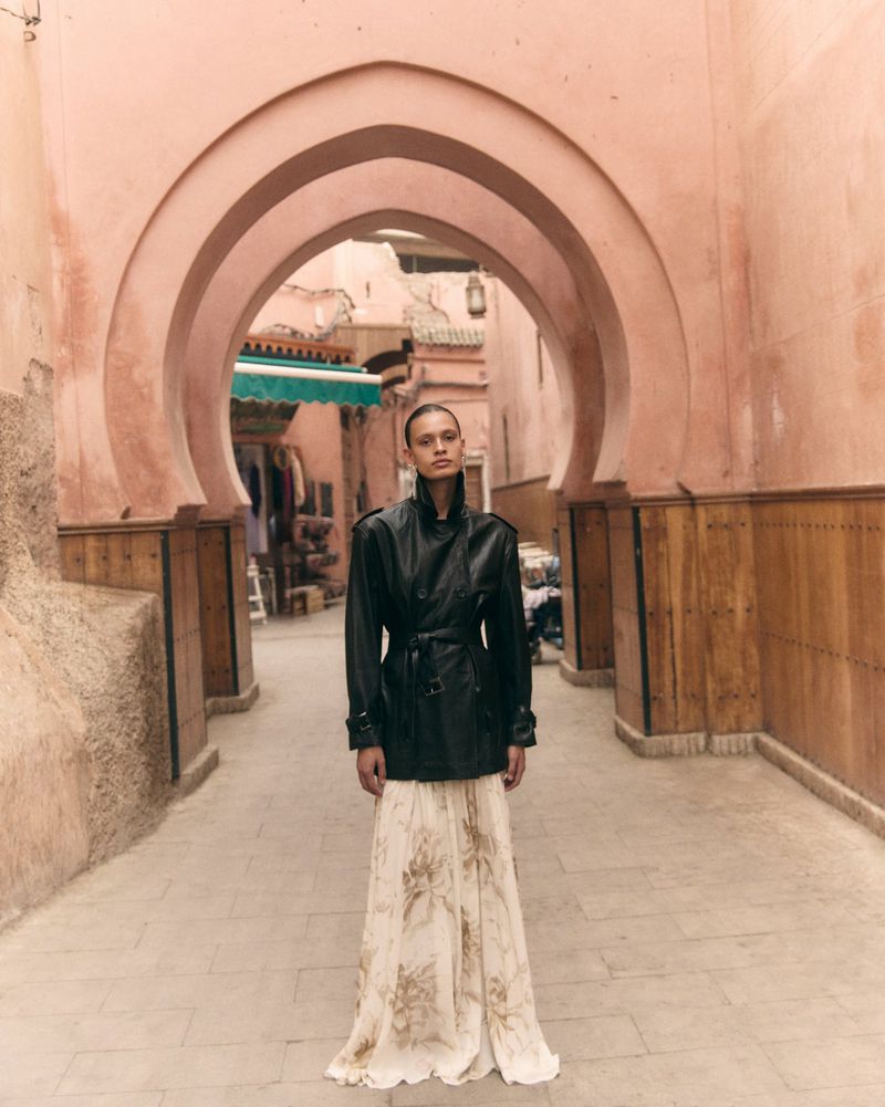Model wearing a belted black leather jacket and flowing printed maxi skirt standing beneath a terracotta archway in an alley.