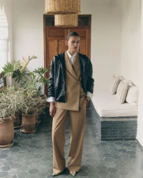 Model wearing a tan tailored suit layered under a black hooded jacket, standing on a hexagonal tiled floor by terracotta planters.