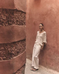 Model in an off-white lace top and matching wide-leg trousers leaning against a textured terracotta wall in a narrow alleyway.