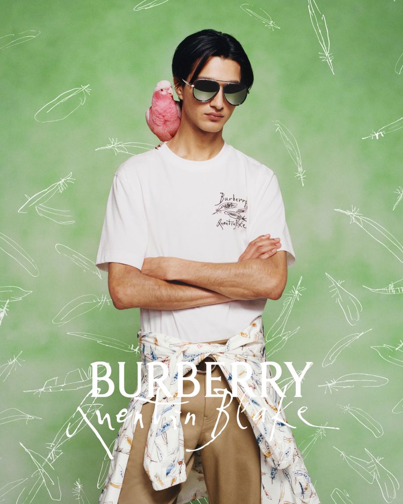 Model wearing dark sunglasses and a white t-shirt with a pink bird on his shoulder against a green illustrated background