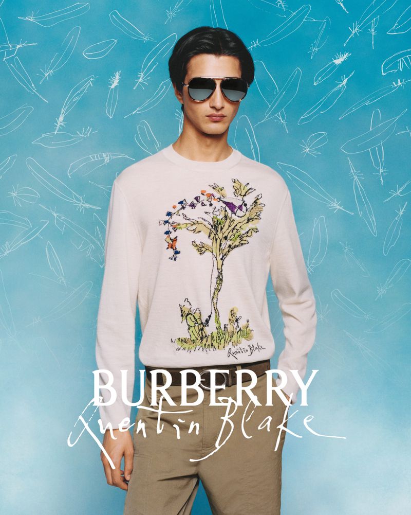 Model Rubuen Bilan-Carroll wearing an illustrated knit sweater and aviator sunglasses against a light blue feather background.