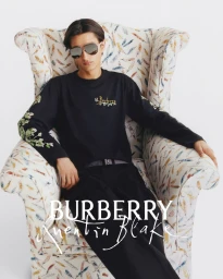 Model Rubuen Bilan-Carroll seated in a feather-patterned armchair wearing a black embroidered long-sleeve top and sunglasses.