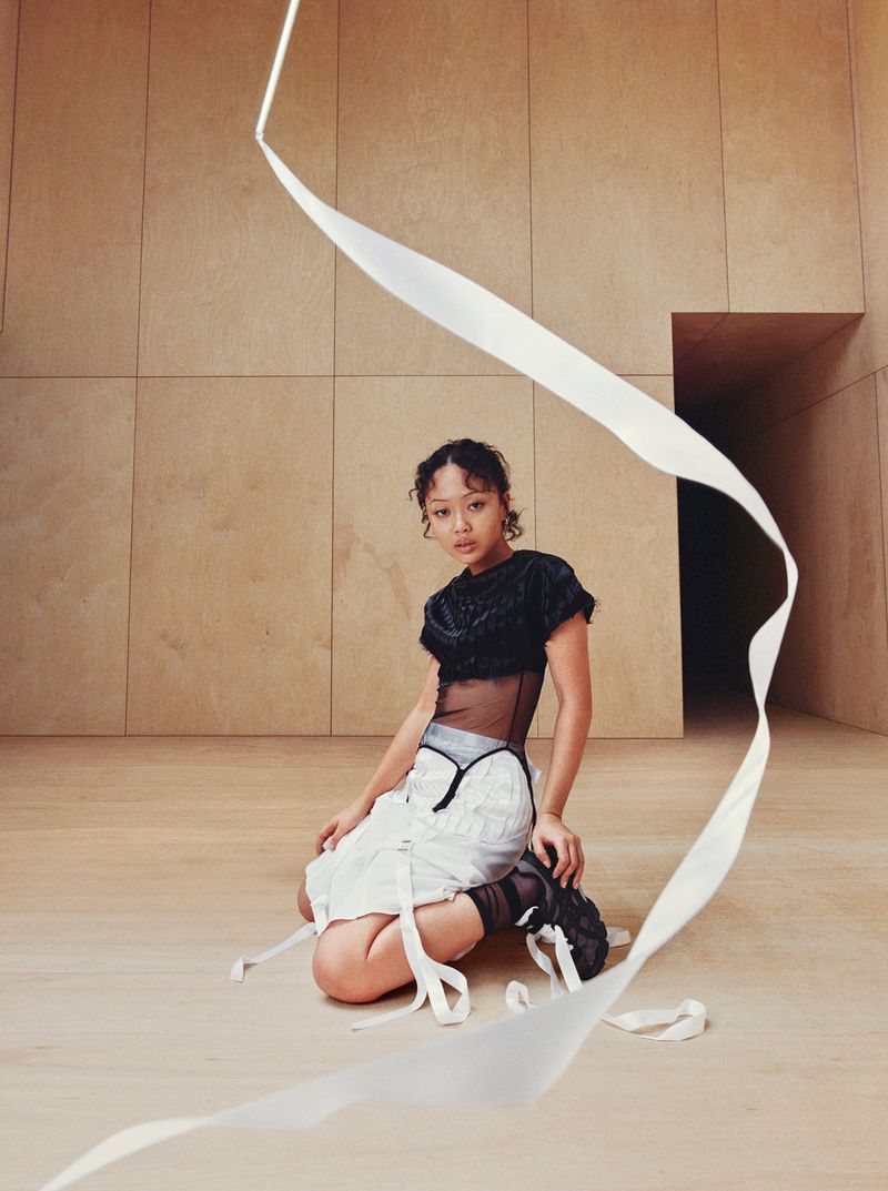 Model kneeling on a wood floor in a modern paneled room with a flowing white ribbon swirling in the foreground.