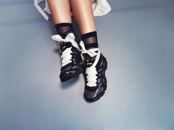 Overhead view of legs wearing black ASICS sneakers with oversized white ribbon bow laces and sheer black socks.