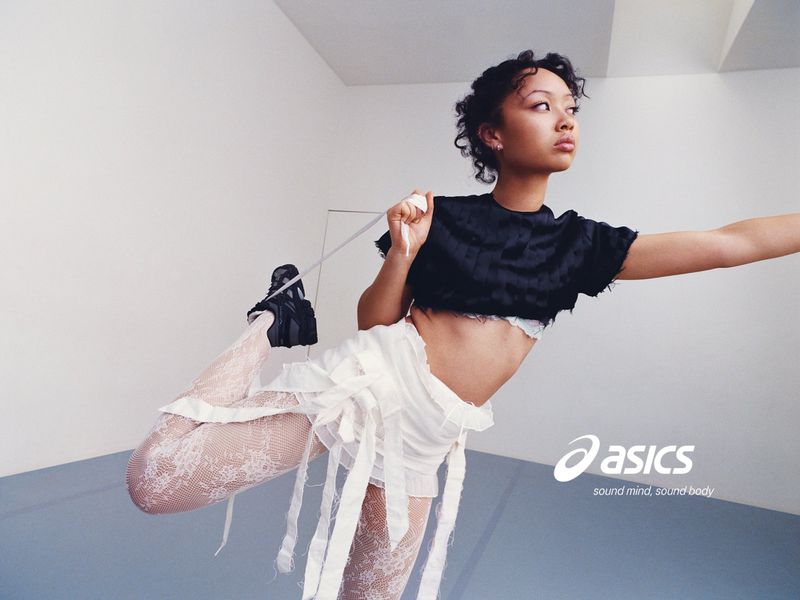 Griff poses dynamically on one leg wearing a black cropped top, white ribbon skirt, lace tights, and black ASICS sneakers in a studio.