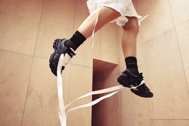 Close-up of legs wearing black sneakers with white ribbons, black socks, and white shorts against wooden paneled wall.