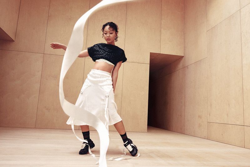Griff poses with a swirling white ribbon wearing a black crop top, white pleated skirt, and black Asics sneakers in a wood-paneled room.