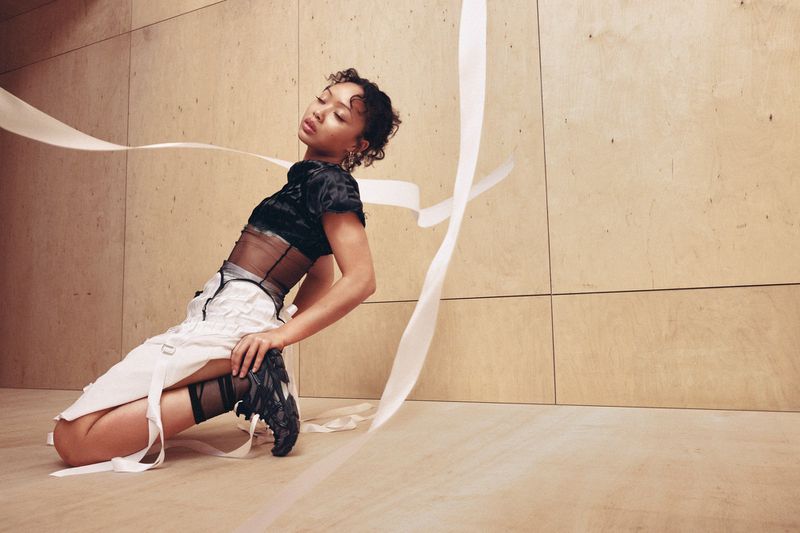 Singer Griff posing dynamically in a black top and white skirt with black ASICS sneakers surrounded by floating white ribbons.