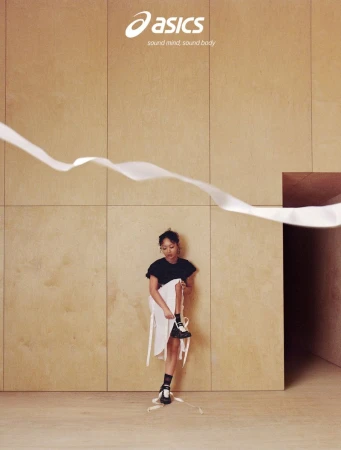 Singer Griff wearing ASICS sneakers, a black top, and a white skirt standing against wooden panel walls with a floating white ribbon.