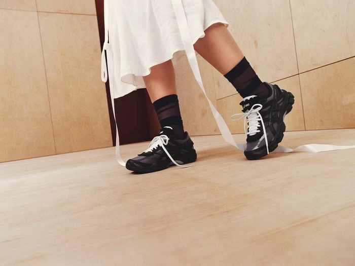 Low-angle shot of a person wearing black ASICS sneakers with white laces, black sheer socks, and a white dress with trailing ribbons.