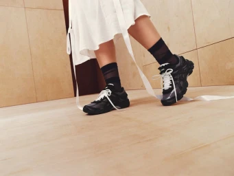 Low-angle shot of a person wearing black ASICS sneakers with white laces, black sheer socks, and a white dress with trailing ribbons.